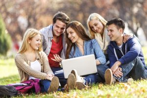 A cheerful group of university students (three girls and two boys) are sitting outdoors in a university campus and having a discussion. The students are using the laptop to surf the Internet and send e-mails. The smiling students are learning and having fun in the same time.